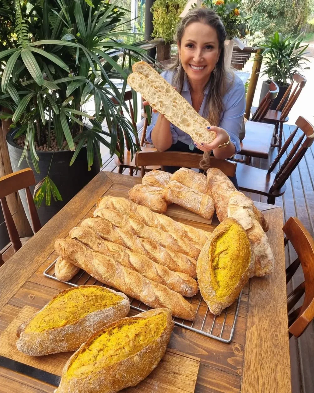 Vivi Lanzellotti na varanda da Casa do Pão na Fazenda, mostrando o miolo de um pão de fermentação natural diante de uma mesa de baguetes e pães italianos