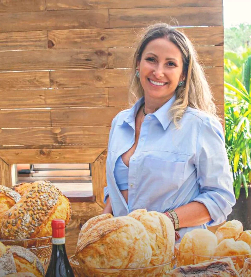 Vivi Lanzellotti, fundadora da Casa do Pão na Fazenda, sorrindo atrás de uma mesa com pães italianos de fermentação natural